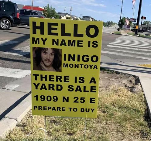 A bright yellow yard‑sale sign staked into grass beside a sidewalk at a street corner. The sign features a photo of Inigo Montoya from The Princess Bride and a parody of his famous line: “Hello, my name is Inigo Montoya. There is a yard sale. 1909 N 25 E. Prepare to buy.”